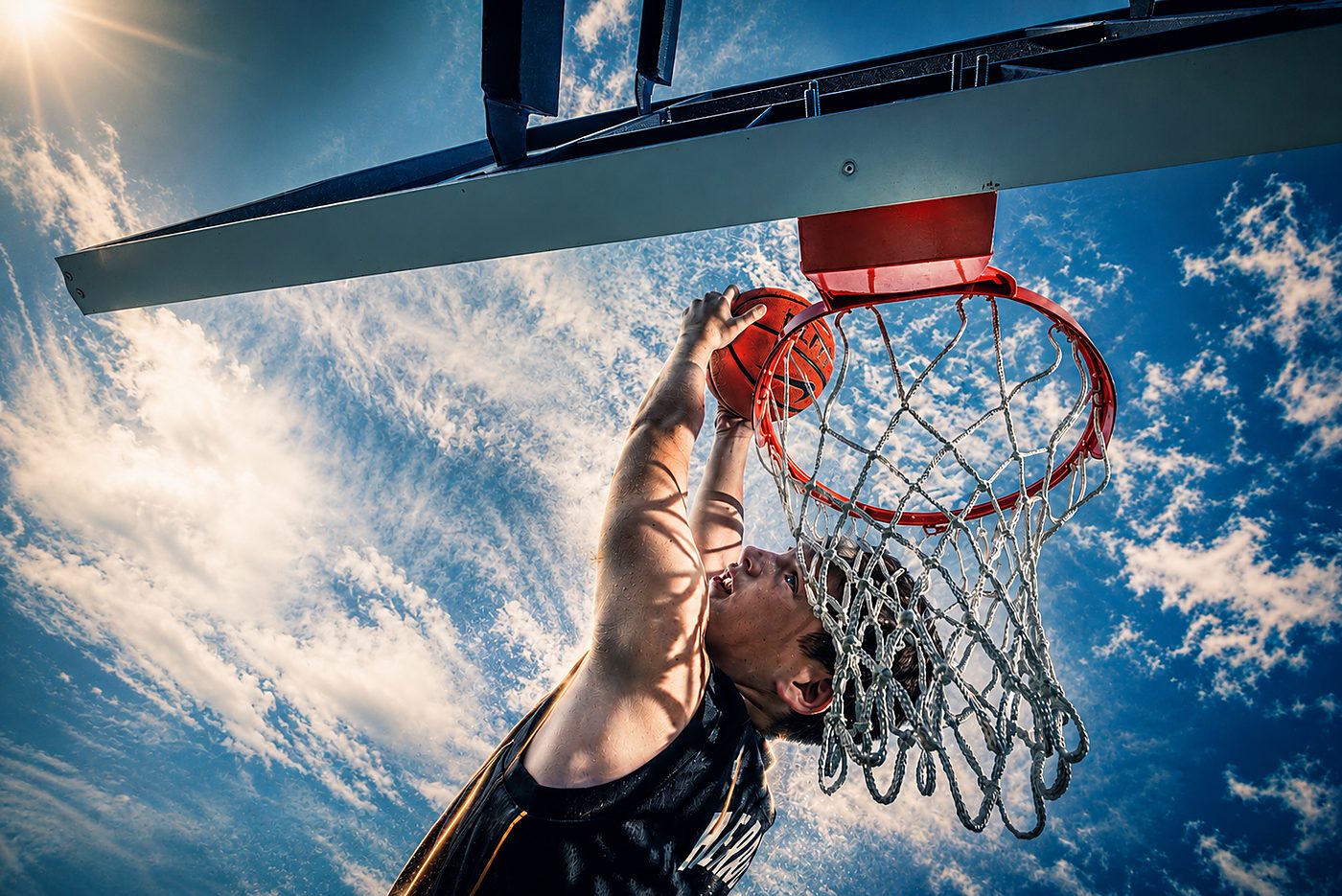 Boy dunking a basketball, seen from below the hoop against a blue sky